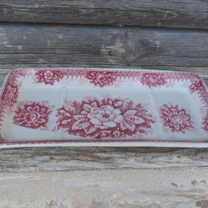 Könnte beinhalten: Ein rechteckiges Serviertablett aus Keramik mit weißem Untergrund und einem dunkelroten Blumenmuster. Das Design zeigt detaillierte Rosenmotive und eine geometrische Bordüre an den Rändern, was diesem Küchenartikel eine klassische Ästhetik verleiht.