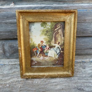 Può includere: Una cornice rettangolare color oro contenente un dipinto di tre figure in un ambiente esterno. La scena raffigura una persona con un abito rosso e blu, un bambino con un vestito rosa e una persona con un vestito bianco vicino a una struttura.