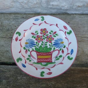 May include: A white ceramic plate with a hand-painted floral design. The design features a pink flower pot with blue and pink flowers and green leaves. The plate has a pink and white border.