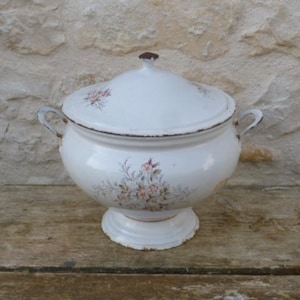 Puede incluir: Sopera de esmalte blanco con tapa, con detalles florales rosas y azules. La sopera tiene una base de pedestal y dos asas laterales. El borde y el pomo de la tapa son marrones. Sobre una superficie de madera.