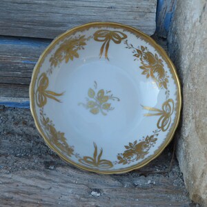 May include: A white porcelain bowl with a gold rim and intricate gold floral and bow designs. The bowl is perfect for serving desserts or snacks.