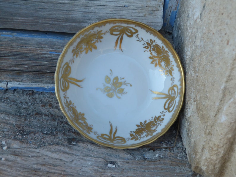 May include: A white porcelain bowl with a gold rim and intricate gold floral and bow designs. The bowl is perfect for serving desserts or snacks.