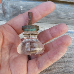 Può includere: Un pomello di vetro trasparente con una base bulbosa e uno stelo filettato in metallo arrugginito. Il pomello ha una piccola parte superiore rotonda con un accento verde. Il pomello è probabilmente per un armadio o un cassetto.