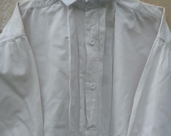 May include: A white long-sleeved shirt with a button-down front and a pleated bib. The shirt has a classic collar and is hanging on a vintage hanger.
