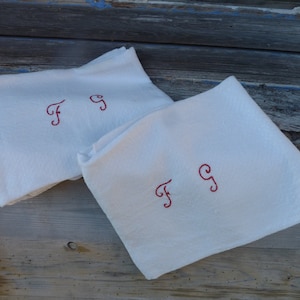 Pode incluir: Duas toalhas de chá de linho branco com iniciais bordadas em vermelho "FG" em cada uma.