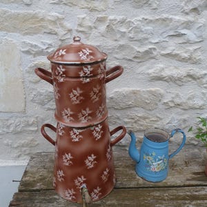 May include: A vintage brown enamel coffee pot with a floral pattern and a small blue pitcher with floral decals. The coffee pot has a spigot and two handles. The items are on a weathered wooden surface.