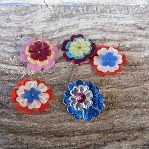 Könnte beinhalten: Fünf farbenfrohe Blumenbroschen mit geschichteten Blütenblättern und einem zentralen Schmuckstein. Die Broschen zeigen eine Vielzahl von Farben, darunter Rosa, Rot, Blau und Gelb. Sie sind auf einer strukturierten Holzoberfläche angeordnet.
