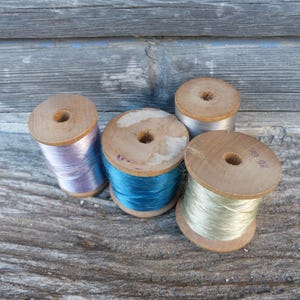 Può includere: Quattro rocchetti di filo di legno vintage nei colori lavanda, turchese, argento e verde chiaro. I rocchetti sono disposti su una superficie di legno grigio e invecchiato, che mostra la venatura del legno.