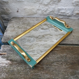 Op de afbeelding: Rechthoekige spiegel met een turquoise lijst en gouden rand. De spiegel heeft gouden handgrepen en decoratieve accenten. De spiegel ligt op een verweerd houten oppervlak.