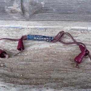 May include: A vintage plum-colored cord with two matching end pieces and a label that reads "POUR VOUS MADAME" in blue and white. The cord is lying on a weathered wooden surface.
