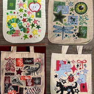 May include: Four beige tote bags with embroidered designs. One has an apple, stars, and a caterpillar. Another features a dragonfly, stars, and an eye. The third has a scorpion, skull, and moth. The last has a cat, stars, and a ladybug.