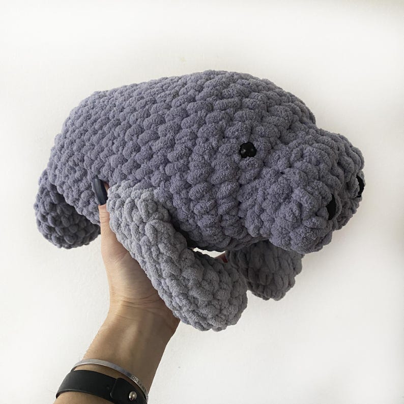 Puede incluir: Un juguete de peluche de manat&iacute; de crochet gris. El juguete est&aacute; siendo sostenido por una mano.