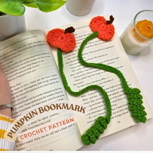 May include: A green crocheted bookmark with two orange crocheted pumpkins attached to the top. The bookmark is laying on top of an open book with the text "Harry Potter and the Philosopher's Stone".