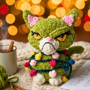 May include: A handmade green crochet cat with a grumpy expression, pink inner ears, and yellow eyes. The cat is wrapped in a string of colourful Christmas lights. A mug of hot chocolate and a ball of yarn are in the background.