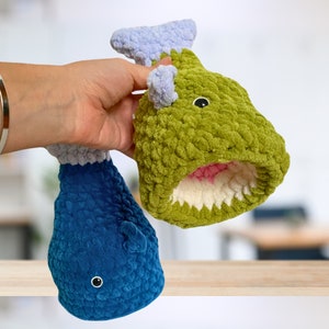 Puede incluir: Dos juguetes de peluche de pescado tejidos a crochet. Uno es verde con una aleta azul claro y el otro es azul con una aleta gris.