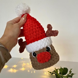 May include: A handmade, brown crocheted reindeer with a red Santa hat, white trim, and a white pom-pom. The reindeer has brown antlers, a red nose, and black eyes. The item is held by a person.