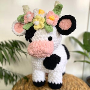 May include: A handmade crochet cow with a white body, black spots, and a pink nose. The cow has a floral crown with pink, yellow, and white flowers. The ears are black with pink insides. The cow is a soft, plush toy.