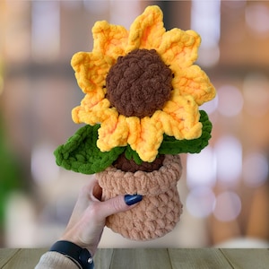 Puede incluir: Un girasol de crochet con pétalos amarillos y un centro marrón, en una maceta de crochet de color beige. El girasol es una artesanía hecha a mano.