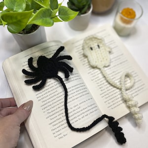 May include: Two crocheted bookmarks, one black spider and one white ghost, are placed on an open book. The spider bookmark has a long tail and eight legs. The ghost bookmark has a long tail and a rounded head.