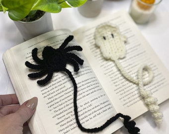 May include: Two crocheted bookmarks, one black spider and one white ghost, are placed on an open book. The spider bookmark has a long tail and eight legs. The ghost bookmark has a long tail and a rounded head.