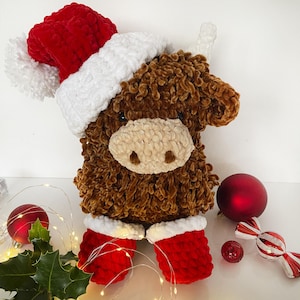 May include: A plush brown Highland cow wearing a red and white Santa hat and red boots with white trim. The stuffed animal has a light beige muzzle and small black eyes. Christmas ornaments and lights are in the background.