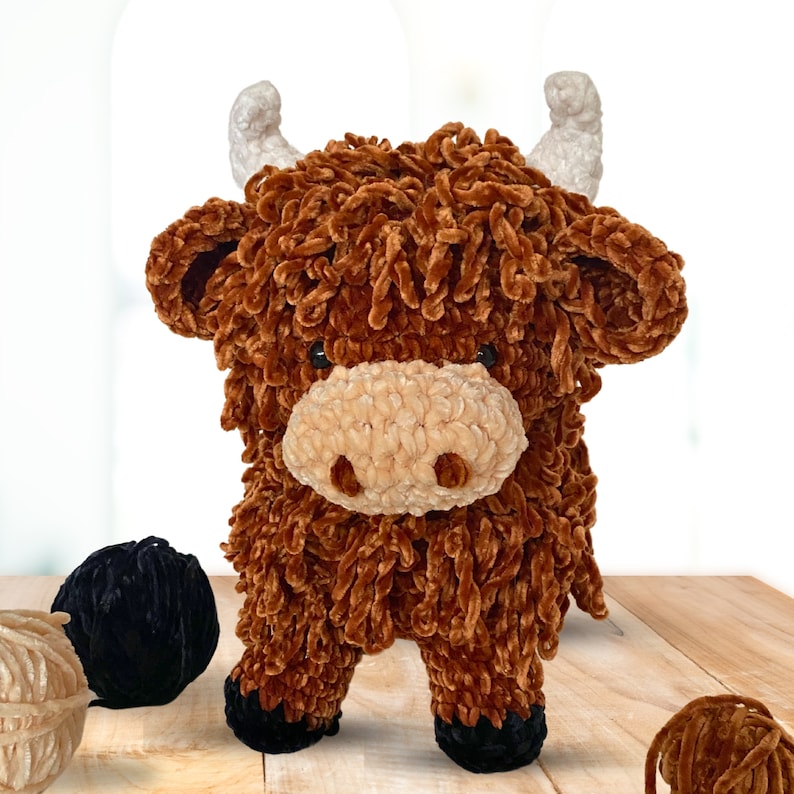 May include: A brown and cream crocheted stuffed animal resembling a Highland cow with long, shaggy fur. The toy has a fluffy tail and large, brown eyes.