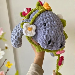 Puede incluir: Una cabeza de burro hecha a mano, de felpa, de color lavanda, con una corona floral y detalles de vid verde. Los ojos y la nariz del burro son negros. Las flores son blancas, rosas y amarillas. El burro está hecho de un hilo suave y texturizado.