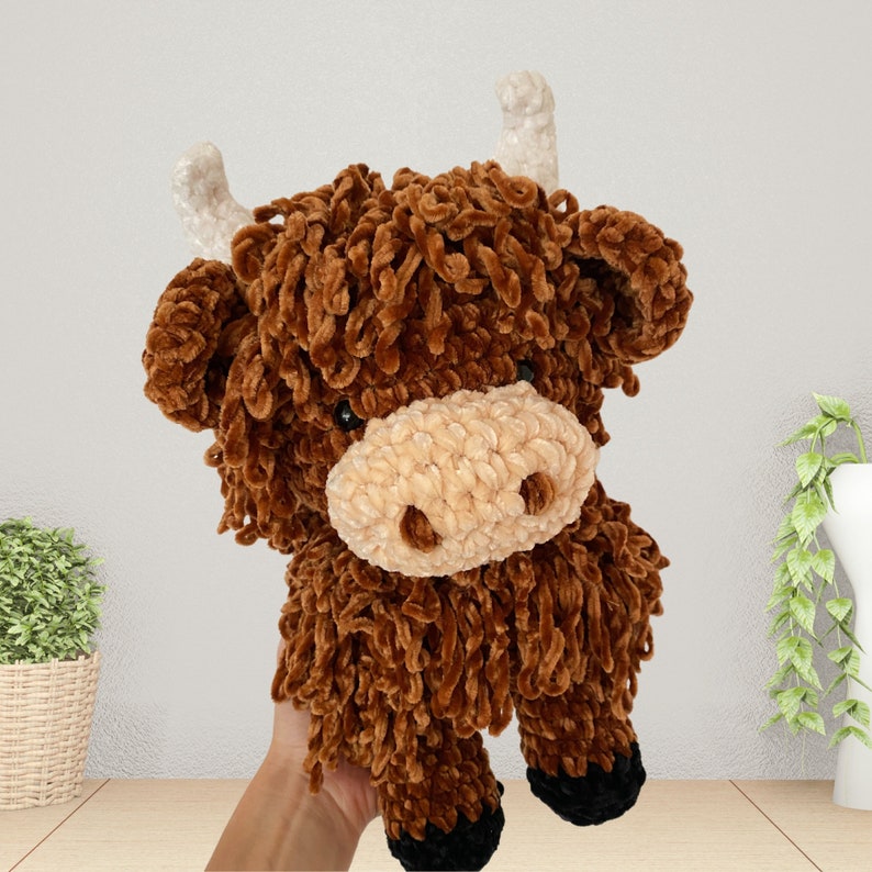 May include: A brown and white crocheted stuffed highland cow toy with a fluffy texture. The toy has a long, curly brown mane and a white face with a brown nose.