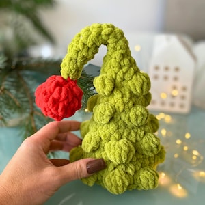 May include: A handmade, green crocheted Christmas tree with a red pom-pom ornament. The tree has a whimsical, textured design and is held by a person's hand. The tree is a festive decoration.