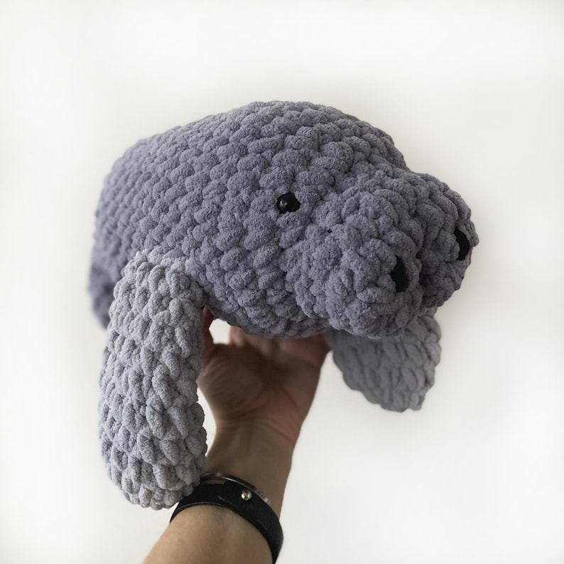 Puede incluir: Un juguete de peluche de manat&iacute; de crochet gris con ojos negros. El juguete est&aacute; siendo sostenido por una mano.