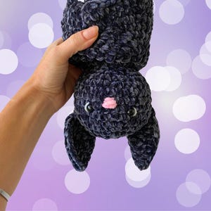 May include: A plush crocheted bat toy with black velvet fabric, black eyes, and a pink nose. The bat is hanging upside down.
