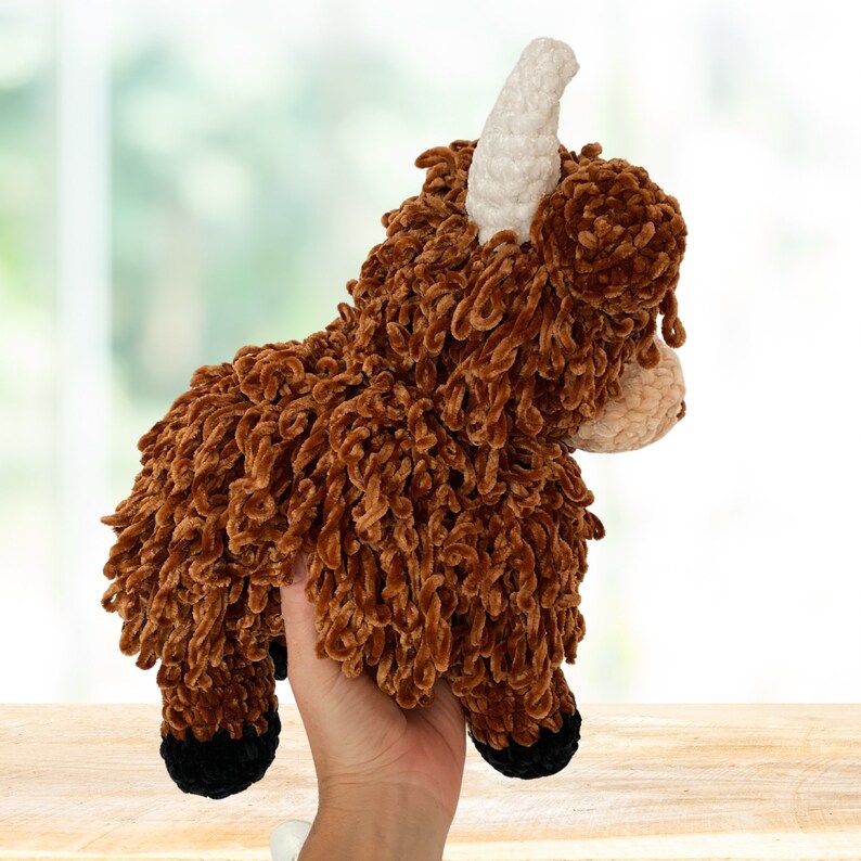 May include: A brown and white crocheted stuffed animal resembling a Highland cow with a fluffy texture. The animal has a white horn and black hooves.