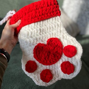 May include: White crocheted stocking with a red heart and paw print design. The stocking has a red crocheted cuff.