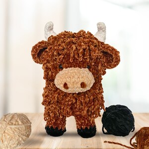 May include: A brown and white crocheted stuffed highland cow toy with a fluffy texture. The toy is standing on a wooden surface with yarn balls in the background.