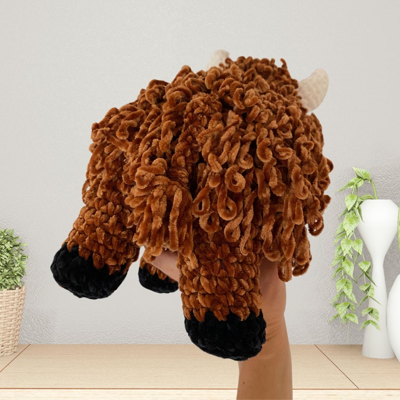 May include: A brown and black crocheted stuffed animal resembling a Highland cow. The animal has a curly, shaggy fur texture and black hooves.