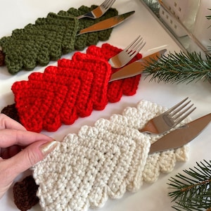 Puede incluir: Soportes de cubiertos con forma de árbol de Navidad de ganchillo en verde, rojo y blanco, cada uno con un tenedor y un cuchillo. Los soportes están sobre una superficie blanca. Decoración festiva.