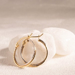 May include: A pair of gold and silver hoop earrings. The earrings are oval-shaped with a textured surface. The earrings are displayed on a white, textured surface. The earrings have a gold clasp.