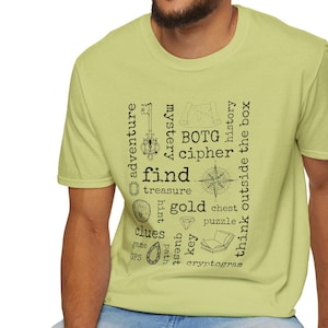 May include: A light green t-shirt with black text and graphics related to treasure hunting. The shirt features words like "adventure," "find," "gold," and "clues," along with a compass and other symbols. The shirt is worn by a person.