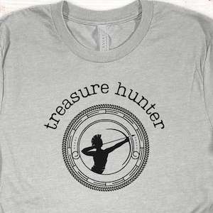 May include: A light gray t-shirt with the words "treasure hunter" in a circular design. The design features a black silhouette of a person with a bow and arrow. The shirt is made of a soft material.