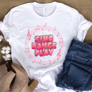 May include: A white short-sleeved t-shirt featuring a circular graphic of pink musical notes and staves surrounding the text SING DANCE PLAY in a bold, gradient pink and orange bubble font with a subtle dotted texture effect.