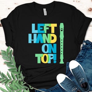 May include: A black short-sleeved t-shirt featuring the text "LEFT HAND ON TOP!" in layered blue, green, and yellow block letters. To the right of the text is a vertical graphic of a light green recorder musical instrument.