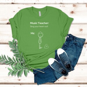 May include: A lime green t-shirt featuring the text "Music Teacher: Sing your heart out! Me:" and a stick figure illustration. The shirt is styled with blue denim jeans and navy trainers with white soles.