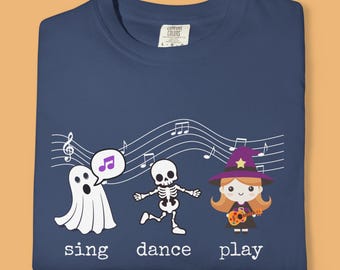May include: Navy blue t-shirt with a musical Halloween theme. Features a ghost, skeleton, and witch with musical notes and the words "sing dance play". The shirt is made of a soft material.