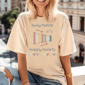May include: A light beige t-shirt with the text "busy hands happy hearts" and a colorful xylophone graphic. The graphic includes musical notes and hearts.