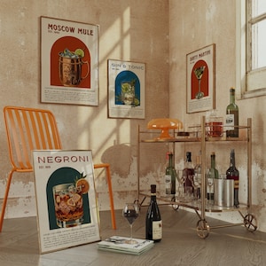 May include: A gold bar cart with bottles of liquor and glasses. There are four vintage posters on the wall behind the bar cart. The posters are for Moscow Mule, Gin & Tonic, Dirty Martini, and Negroni. The posters are all in a retro style with illustrations of the drinks.