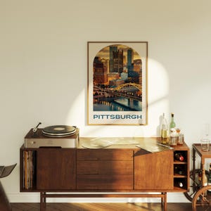 K&ouml;nnte beinhalten: Ein Plattenspieler im Vintage-Stil steht auf einem Holzschr&auml;nkchen mit einem Poster der Skyline von Pittsburgh dar&uuml;ber. Das Poster hat eine Bogenform und das Wort "PITTSBURGH" ist unten in Blau gedruckt.