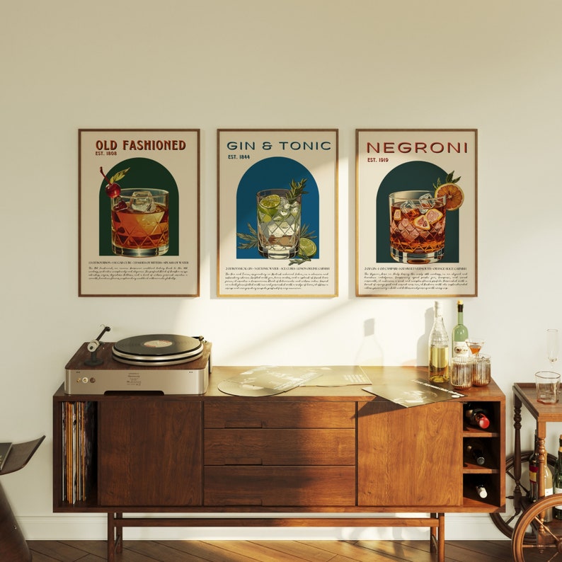 May include: Three vintage-style posters with illustrations of classic cocktails. The posters are framed and hung on a white wall above a wooden cabinet with a record player on top. The posters feature the names "Old Fashioned", "Gin & Tonic", and "Negroni" with the year they were established.