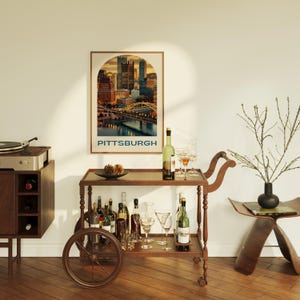 K&ouml;nnte beinhalten: Ein Barwagen aus Holz mit einer braunen Oberseite und R&auml;dern. Der Wagen ist gef&uuml;llt mit Weinflaschen, Gl&auml;sern und einer dekorativen Kiefernzapfen-Anordnung. Ein Poster mit einer Stadtansicht und dem Wort "PITTSBURGH" h&auml;ngt an der Wand hinter dem Wagen.