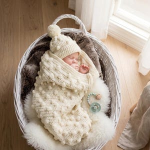 May include: A sleeping baby wrapped in a cream-colored, textured knit blanket and hat with a pom-pom, nestled in a white wicker basket. The basket sits on a fluffy white rug. A baby mobile hangs above. A pacifier rests nearby.