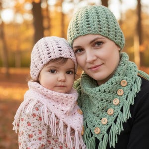 May include: A mother and child wearing matching crocheted hats and scarves. The mother's scarf is sage green with wooden buttons and fringe. The child's hat and scarf are light pink. The child is wearing a floral print dress.